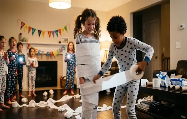 A group of young kids in pajamas playing a toilet paper mummy wrap relay race game in a cozy living room decorated with a Back to School banner.