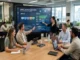 A modern marketing operations team sitting around a conference table reviewing a large digital display showing campaign workflows, resource allocation, and budget analytics.