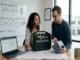 Two product developers sitting at a desk with a portable power station, technical blueprints, and a laptop spreadsheet, in front of a whiteboard explaining OEM vs ODM pathways.