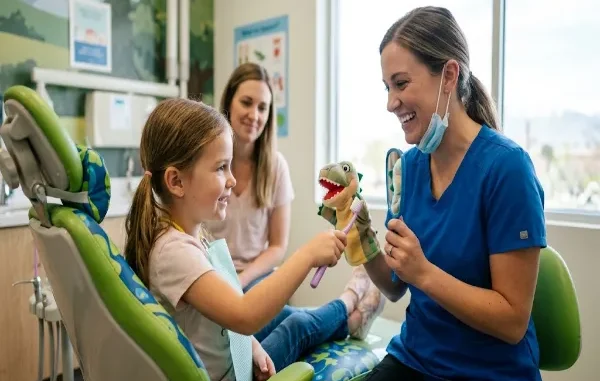 A friendly pediatric dentist smiling while showing a young girl in a dental chair how to brush a puppet's teeth, as the girl's mother watches in the background.
