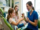 A friendly pediatric dentist smiling while showing a young girl in a dental chair how to brush a puppet's teeth, as the girl's mother watches in the background.