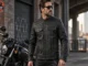 A male rider wears a black leather motorcycle jacket with distinct white stitching, showcasing a panel with a hexagonal quilted or perforated pattern on the chest and shoulders, with a cruiser motorcycle in the background.