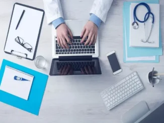 An overhead, flat-lay photograph shows the hands of a medical professional in a white lab coat typing on an open laptop positioned centrally on a light gray wooden desk. Surrounding the laptop are various administrative and medical items, including a clipboard with papers and glasses, a stethoscope on a blue folder, a spiral notebook with a pen, a smartphone, a wireless keyboard, a glass of water, and a pen cup.