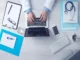 An overhead, flat-lay photograph shows the hands of a medical professional in a white lab coat typing on an open laptop positioned centrally on a light gray wooden desk. Surrounding the laptop are various administrative and medical items, including a clipboard with papers and glasses, a stethoscope on a blue folder, a spiral notebook with a pen, a smartphone, a wireless keyboard, a glass of water, and a pen cup.