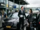 A professional chauffeur in a suit holding a greeting sign and opening a black luxury sedan door for a smiling business traveler outside Amsterdam Airport Schiphol arrivals.