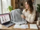 A woman sitting at a wooden desk comparing a grey Raspberry Hills hoodie to an online product page on her laptop while reviewing a size chart and fabric swatch.