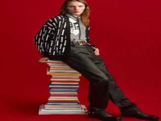Model with long hair leaning against a tall stack of colorful books against a solid red background, wearing a black-and-white patterned cardigan, graphic button-up shirt, gold-buckle belt, black leather trousers, and boots.