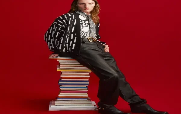 Model with long hair leaning against a tall stack of colorful books against a solid red background, wearing a black-and-white patterned cardigan, graphic button-up shirt, gold-buckle belt, black leather trousers, and boots.