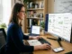 A side-view photograph of a woman wearing glasses and a blue top, sitting at a wooden desk in a home office and working on a computer. Her large monitor displays a "Centralised Reputation Dashboard," showing data from Google, Yelp, and Trustpilot. A notebook, keyboard, mouse, and phone are on the desk, with a bookcase and whiteboard in the background.
