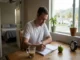 A man sitting at a wooden kitchen table writing in a daily planner next to a green apple, alarm clock, and a cup of coffee.