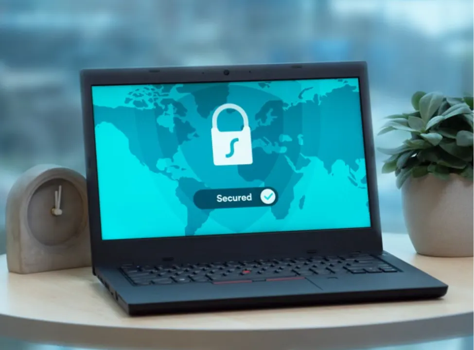 Black laptop open on a table displaying a teal Surfshark VPN shield logo, world map, and a button indicating Secured with a checkmark.
