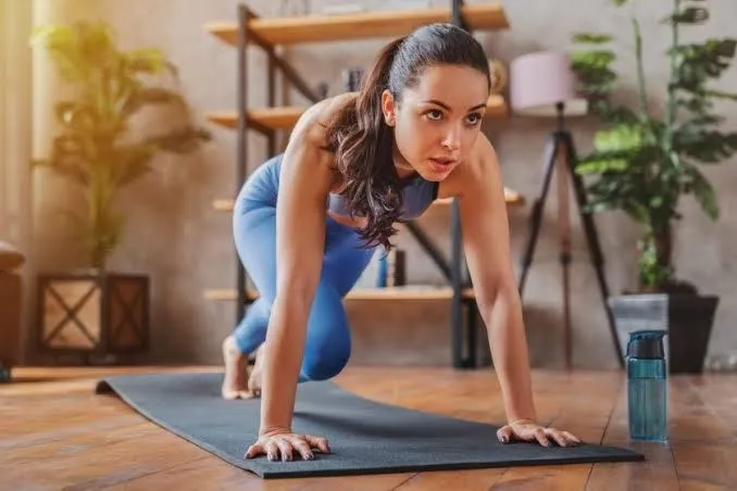 A young woman in a blue sports top and leggings is focused and performing a core-strengthening exercise. She is in a plank position with her arms straight, on a dark mat on a wooden floor, and one knee is tucked towards her chest. The bright, loft-like room features potted plants and wooden shelving in the background.