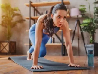 A focused young woman in blue athletic wear performing plank knee drives on a black yoga mat in a sunlit room.