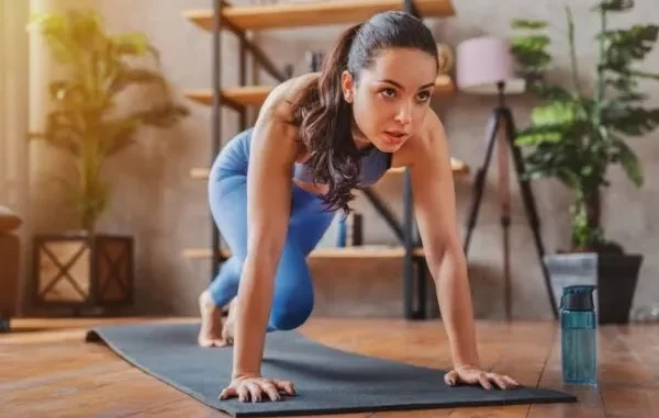A focused young woman in blue athletic wear performing plank knee drives on a black yoga mat in a sunlit room.