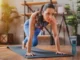 A focused young woman in blue athletic wear performing plank knee drives on a black yoga mat in a sunlit room.