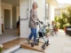 An active senior woman with short grey hair smiling while using a modern blue rollator walker to cross a low-threshold wood ramp leading from her open doorway onto a sunny patio.