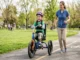 A young girl with leg braces smiling while riding a bright blue adaptive tricycle in a park, accompanied by a female physical therapist running alongside her on a paved path.