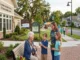 An elderly woman sitting on a park bench outside a modern assisted living community center in Canton, GA, smiling while interacting with two young children and a volunteer in a lively neighborhood setting with walking paths and local shops in the background.