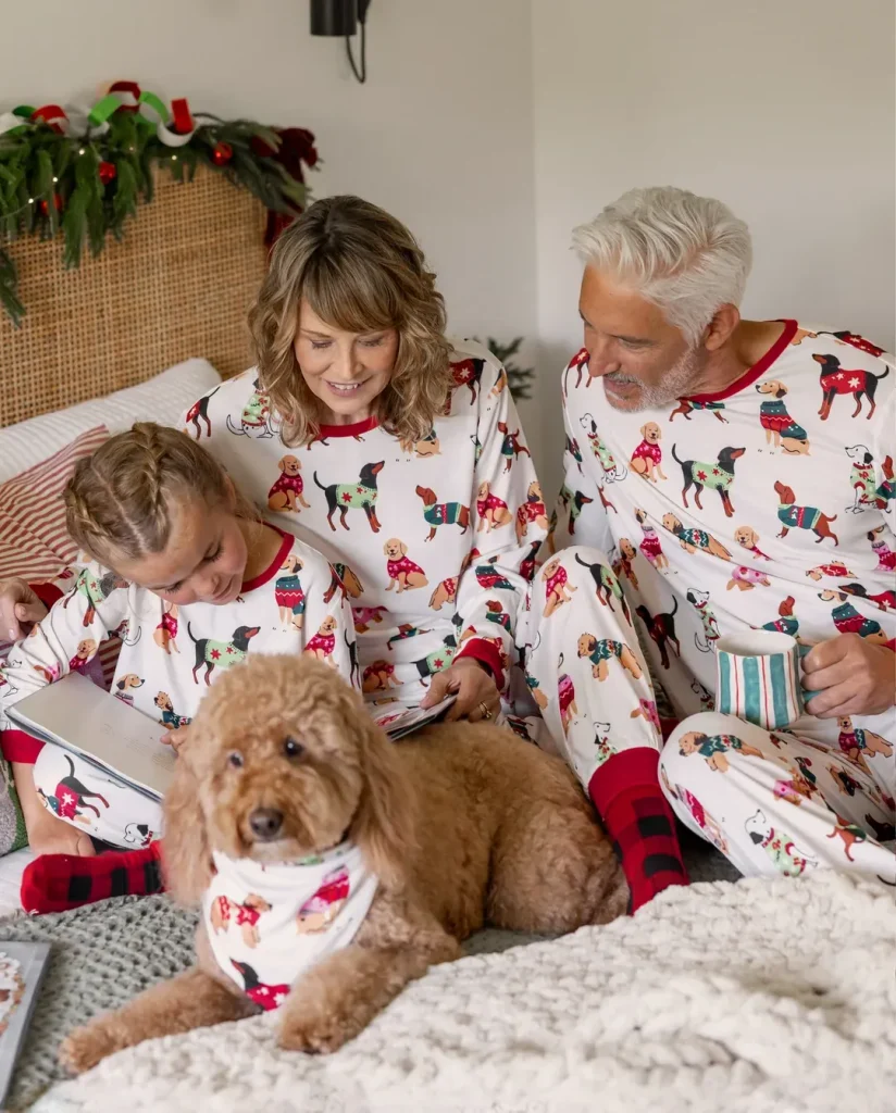 A family sitting together on a guest room bed in matching white holiday pajamas featuring colorful dog patterns, with a fluffy golden dog in the foreground wearing a matching bandana.