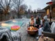 A group of friends sits on outdoor sofas around a blazing copper fire pit, enjoying warm drinks and s'mores by a steaming backyard swimming pool in the evening. A giant donut float is in the water, and everyone is bundled in winter clothes.