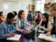 A group of high school students collaborating around a plant growth experiment with grow lights in a modern science classroom as their teacher guides them.