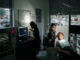 A dark dental clinic during a power outage. One staff member uses a handheld light to examine a patient in a chair while another works at a computer. Rain is visible through the window.