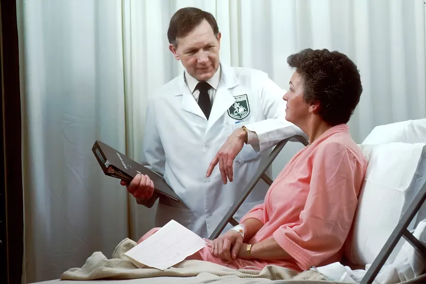 A male doctor in a white coat holds medical charts and speaks to a woman in a pink hospital gown resting in bed with a paper in her hand.