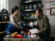 A man and a woman organizing essential emergency preparedness supplies and protective gear, including a blue tarp, heavy-duty gloves, N95 mask, first aid kit, and an inventory checklist.