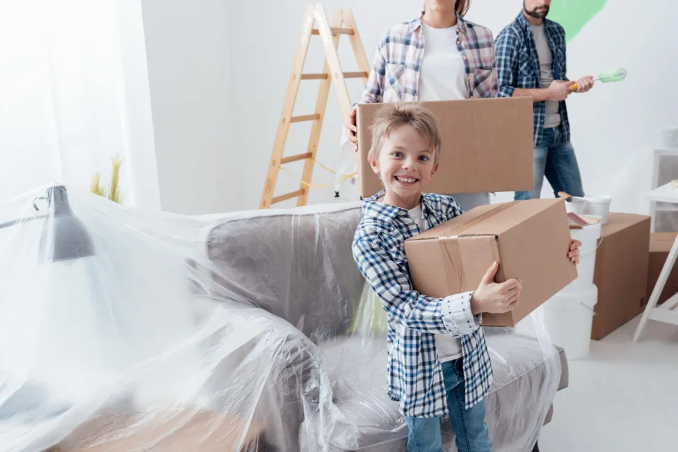 A smiling young boy wearing a blue plaid shirt holding a cardboard moving box in a bright room, with his parents carrying boxes and painting supplies in the background.