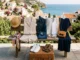 A neatly arranged vacation capsule wardrobe displaying a beige linen set, dark denim jeans, striped top, white sneakers, dark navy wrap dress, straw hat, and accessories on a wooden bench against a coastal Mediterranean background.