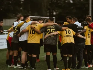 Members of a soccer team standing in a huddle on the pitch, seen from behind wearing yellow, grey, and red jerseys, symbolizing team unity and chemistry in Ultimate Team mode.