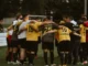 Members of a soccer team standing in a huddle on the pitch, seen from behind wearing yellow, grey, and red jerseys, symbolizing team unity and chemistry in Ultimate Team mode.