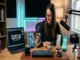 A woman with glasses and a black sweater sits at a large wooden desk surrounded by multiple gadgets, including a laptop with a tech review title, smartphones on stands, a camera, a keyboard, and a microphone.