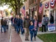 A multi-generational line of community members and veterans walking down a brick sidewalk decorated with American flags toward a "Vote Here" polling place.