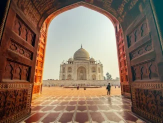 View through a large, ornate carved wooden archway towards the white marble Taj Mahal and its dome in Agra, India, with a person walking on the red sandstone courtyard.