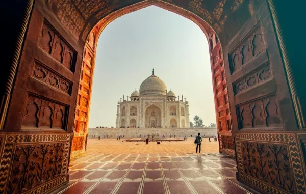 View through a large, ornate carved wooden archway towards the white marble Taj Mahal and its dome in Agra, India, with a person walking on the red sandstone courtyard.