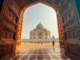 View through a large, ornate carved wooden archway towards the white marble Taj Mahal and its dome in Agra, India, with a person walking on the red sandstone courtyard.
