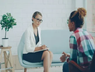 A woman wearing glasses and holding a notepad listens attentively to a client sitting opposite her in a modern office setting.