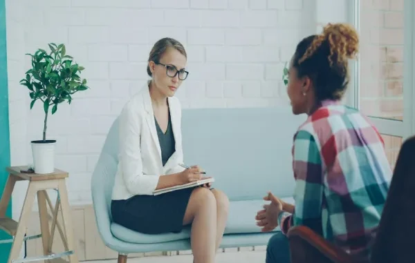 A woman wearing glasses and holding a notepad listens attentively to a client sitting opposite her in a modern office setting.