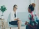 A woman wearing glasses and holding a notepad listens attentively to a client sitting opposite her in a modern office setting.