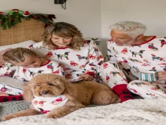A family wearing matching white holiday pajamas with printed dogs and red trim, sitting on a bed reading books with their golden dog wearing a matching bandana.