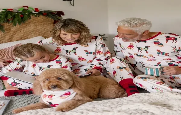 A family wearing matching white holiday pajamas with printed dogs and red trim, sitting on a bed reading books with their golden dog wearing a matching bandana.
