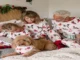 A family wearing matching white holiday pajamas with printed dogs and red trim, sitting on a bed reading books with their golden dog wearing a matching bandana.