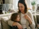 A mother wearing a gold necklace with initial pendants and a charm bracelet sitting on a sofa while hugging her young child.