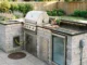 A realistic outdoor kitchen built with stacked stone featuring a stainless steel gas grill, sink, under-counter refrigerator, visible gas line hatch, outdoor electrical conduit, and stone patio drainage grate.