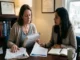 A middle-aged female patient holding medical record files sits across a desk discussing her case with a female attorney in a legal office.
