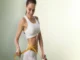 A fit woman in a white sports bra and shorts smiling as she measures her slender waist with a yellow tape measure, representing the potential physical results of peptide therapy.