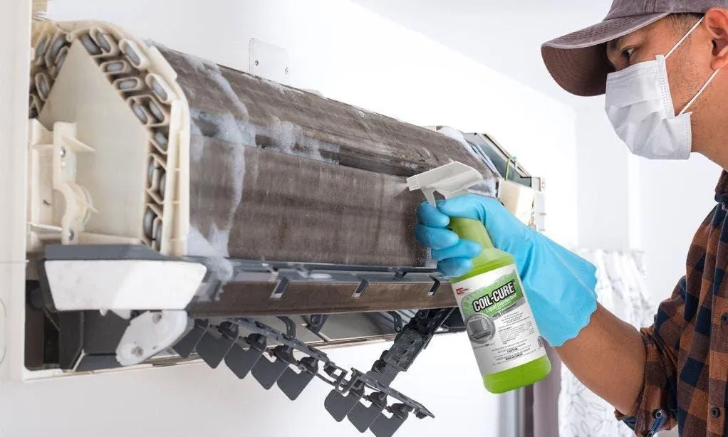 Professional HVAC technician wearing a face mask and protective gloves spraying Coil-Cure disinfectant cleaner onto indoor AC unit coils during deep cleaning maintenance in Dubai.