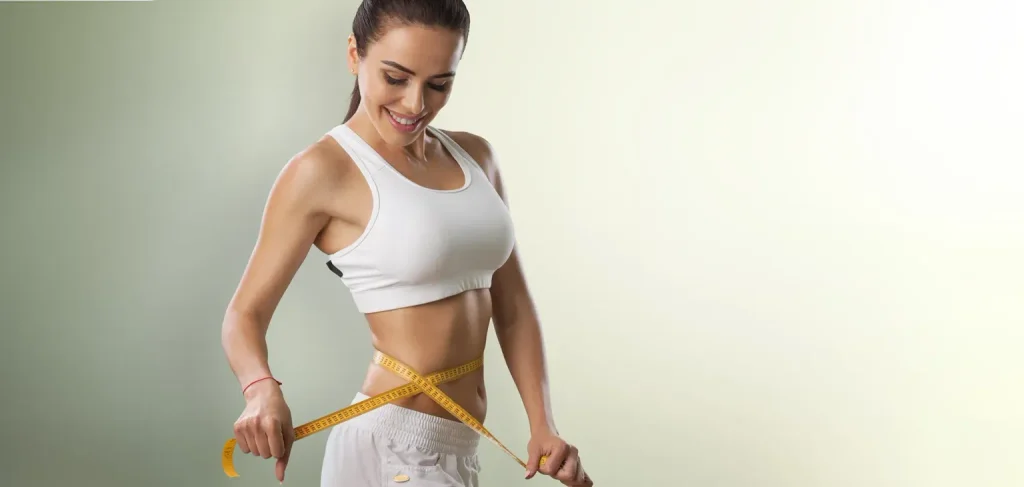 A medium wide-angle shot of a smiling fit young adult woman in a white sports bra and white pants, looking down as she measures her narrow waist with a yellow tape measure, against a light gradient background.