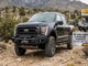 A black Ford off-road truck outfitted with heavy-duty front bumper, winch, skid plate, and rock sliders parked near a 4 Wheel Parts shop.